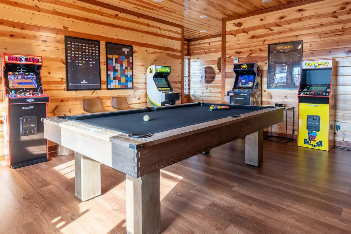 A vibrant game room with a pool table, classic arcade machines, and game-themed artwork on the walls. The space is designed for group fun and friendly competition, with a nostalgic and playful vibe.