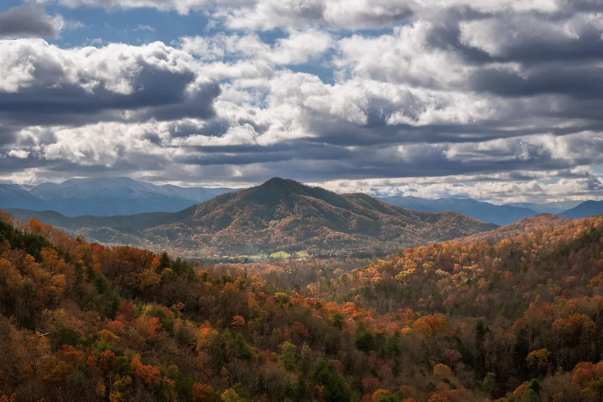 A sweeping view of the Great Smoky Mountains with a cloudy sky and vibrant autumn foliage. The scene evokes the untouched beauty and serenity of the surrounding national park, encouraging thoughts of hiking and outdoor adventure.