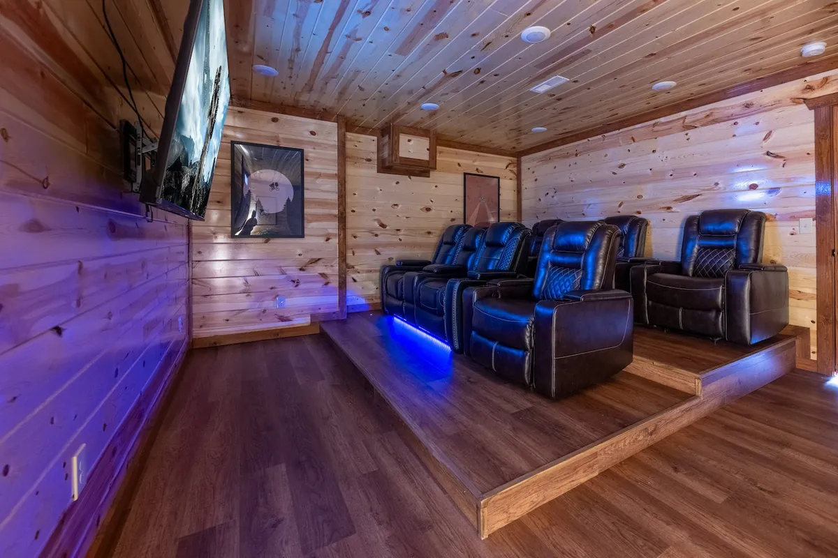 A cozy, immersive private theater room with reclining chairs, a large TV screen, wood-paneled walls, and ambient lighting. The setup suggests comfortable movie nights and shared family experiences.