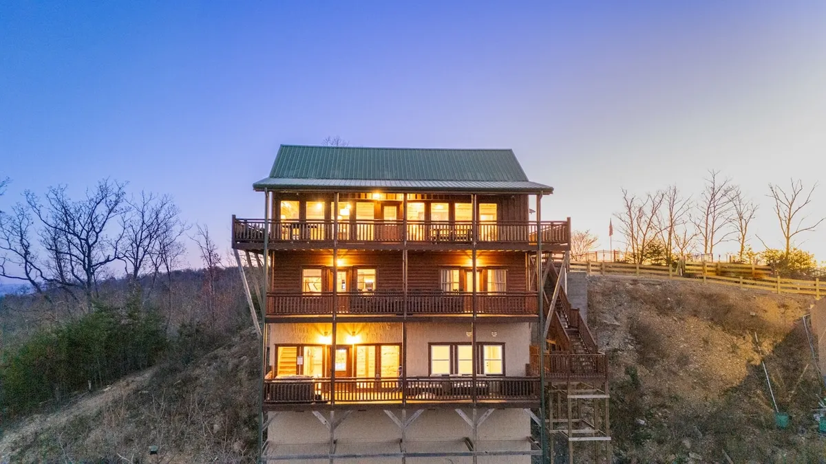 A multi-level cabin with warmly illuminated windows nestled among lush greenery, surrounded by the tranquil beauty of the mountains. The home radiates coziness and hints at the comfort awaiting after a day of exploration.