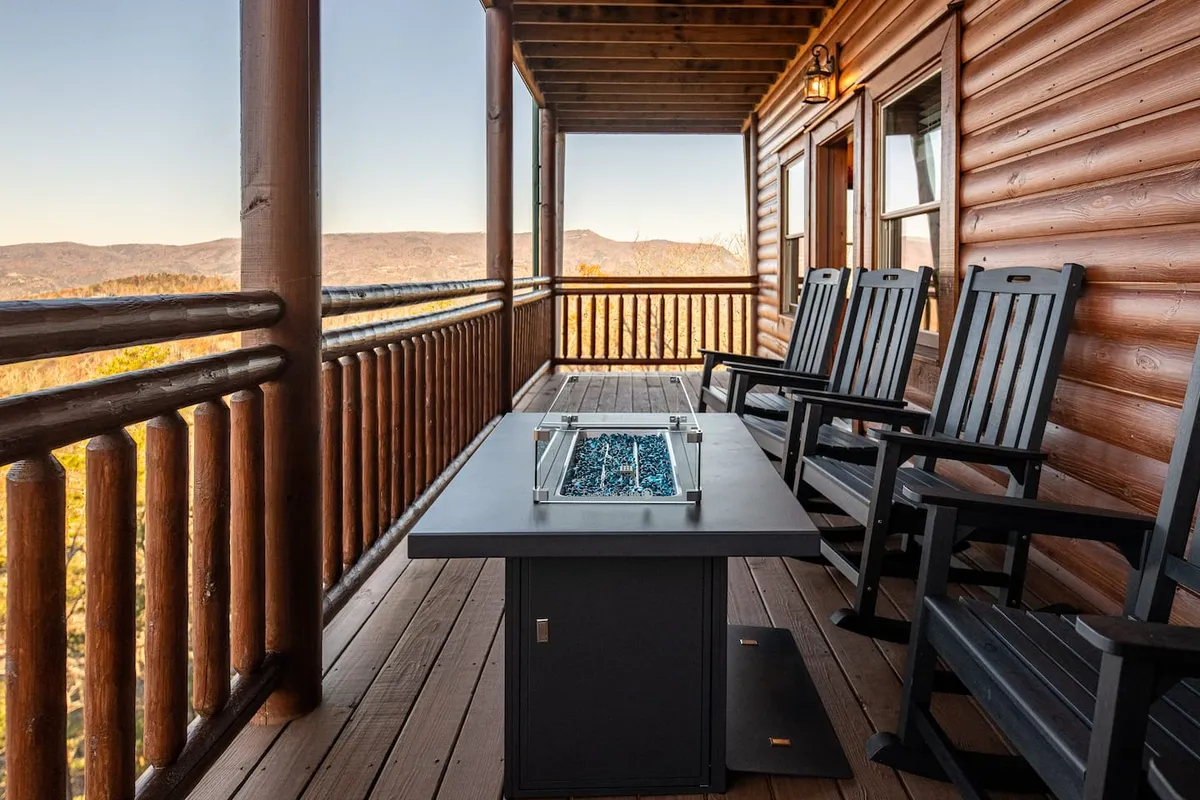 This exterior image features a cozy fire table surrounded by rocking chairs on a wooden deck, with a stunning panoramic mountain view beyond the railing. The inviting setup is clearly designed for groups to gather outdoors, relax, and enjoy both the warmth of the fire and the scenic natural backdrop.