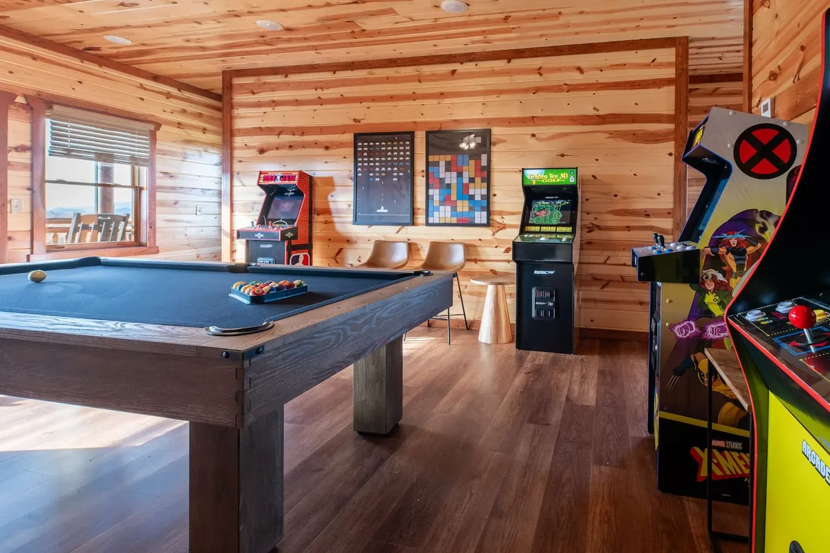 A lively game room featuring a pool table set against rustic wooden walls, flanked by classic arcade machines and artwork on the wall. The space exudes a fun and relaxed vibe, perfectly suited for socializing and friendly competition among groups.
