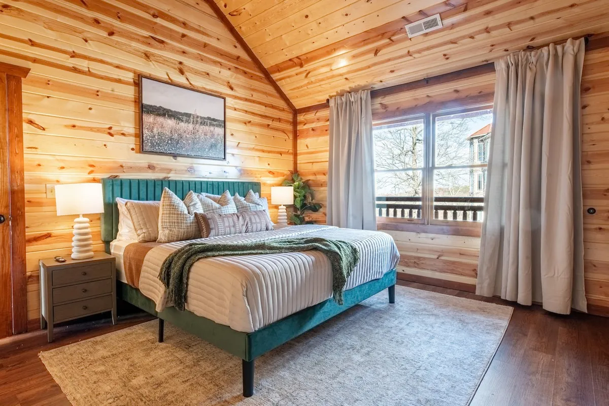 A tranquil bedroom featuring a comfortable bed adorned with decorative pillows and soft bedding, flanked by warm wooden walls. The space is bathed in natural light streaming through a large window, offering a glimpse of the outdoors and creating a peaceful atmosphere for rest and relaxation.