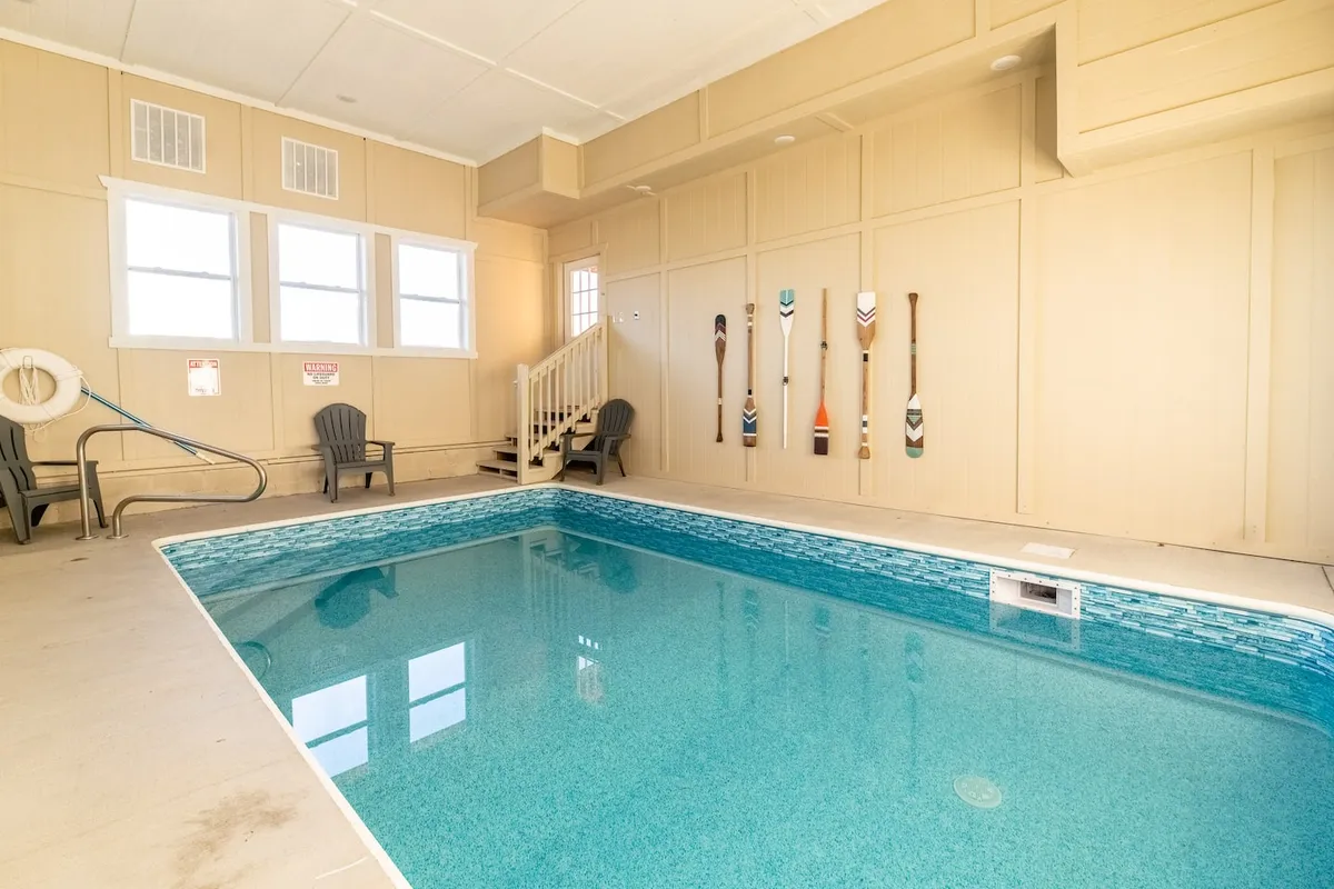 A bright and inviting indoor swimming pool area featuring decorative paddles on the wall and comfortable lounge chairs set along the poolside. The scene captures the tranquil atmosphere, soft lighting, and ample space for relaxation or play.