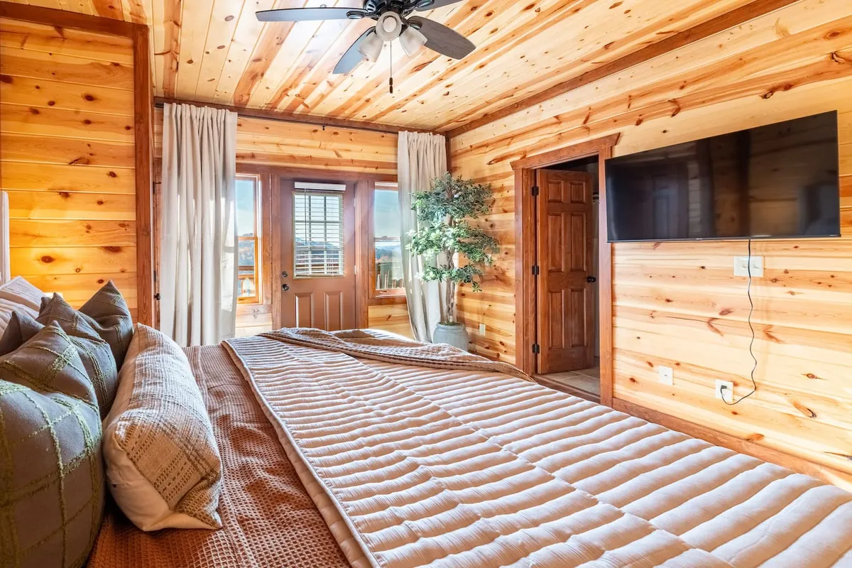 A serene bedroom with a large bed topped with decorative pillows and soft layers, set against wooden walls. A television and an indoor plant add contemporary comfort, while a sunlit window brings in natural brightness—ideal for restful mornings and cozy evenings.