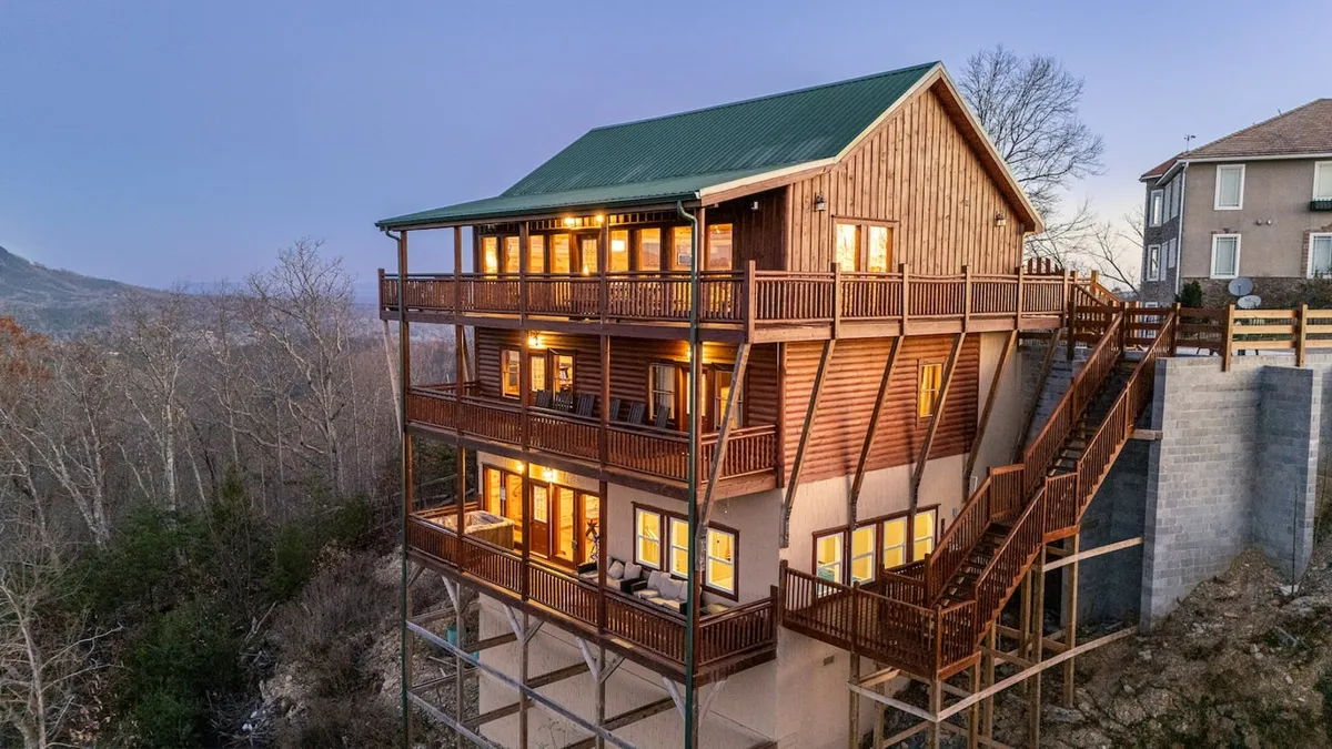 A wide shot of the cabin’s wooden balcony, featuring large windows and a green metal roof, set against a backdrop of lush surrounding nature. The scene evokes a sense of arrival, relaxation, and outdoor enjoyment.