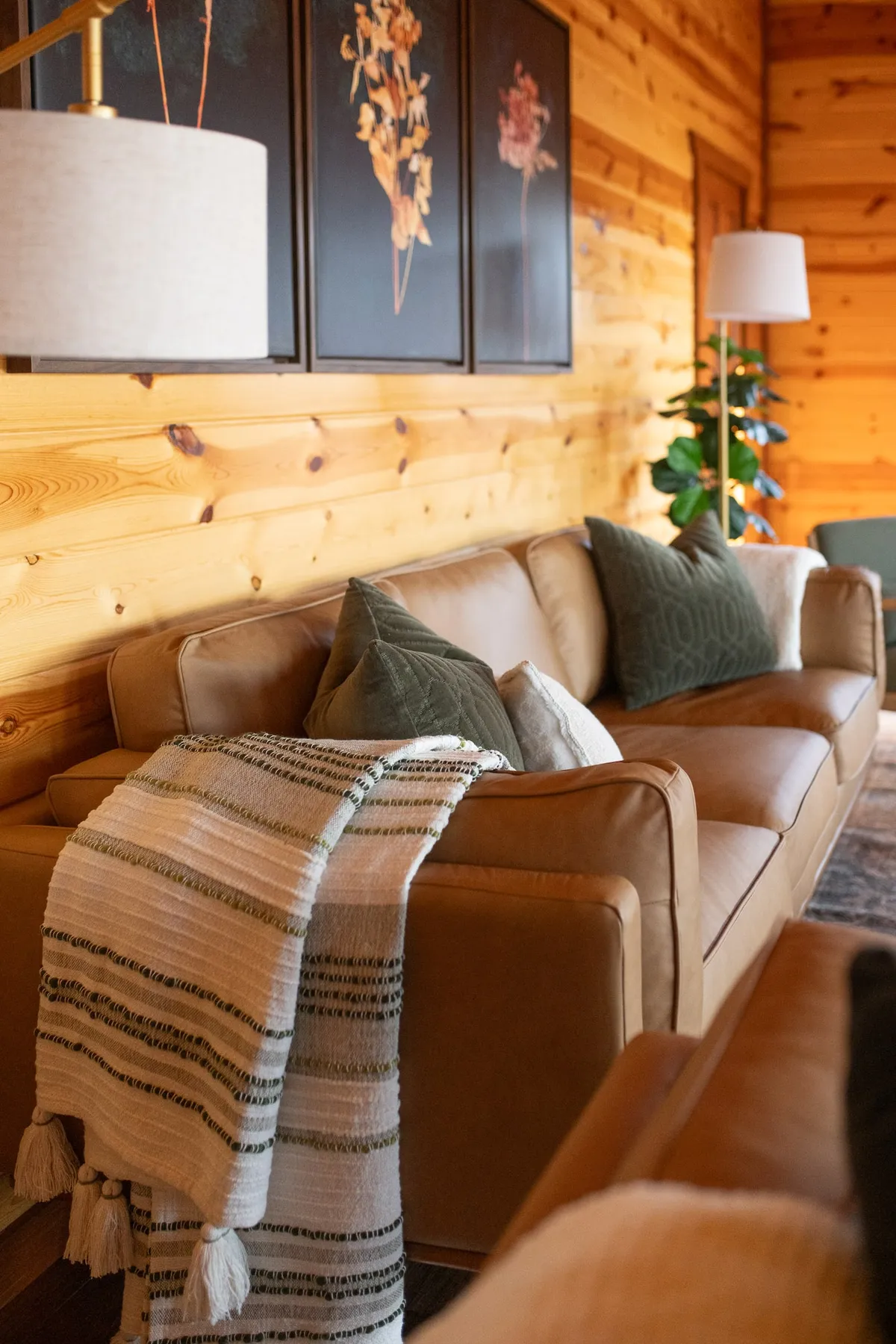 A warm and welcoming living room featuring a leather sofa with decorative pillows, a textured blanket, wall art, a floor lamp, and a lush indoor plant. The setup encourages guests to relax and gather, fostering heartfelt conversations and cozy moments.